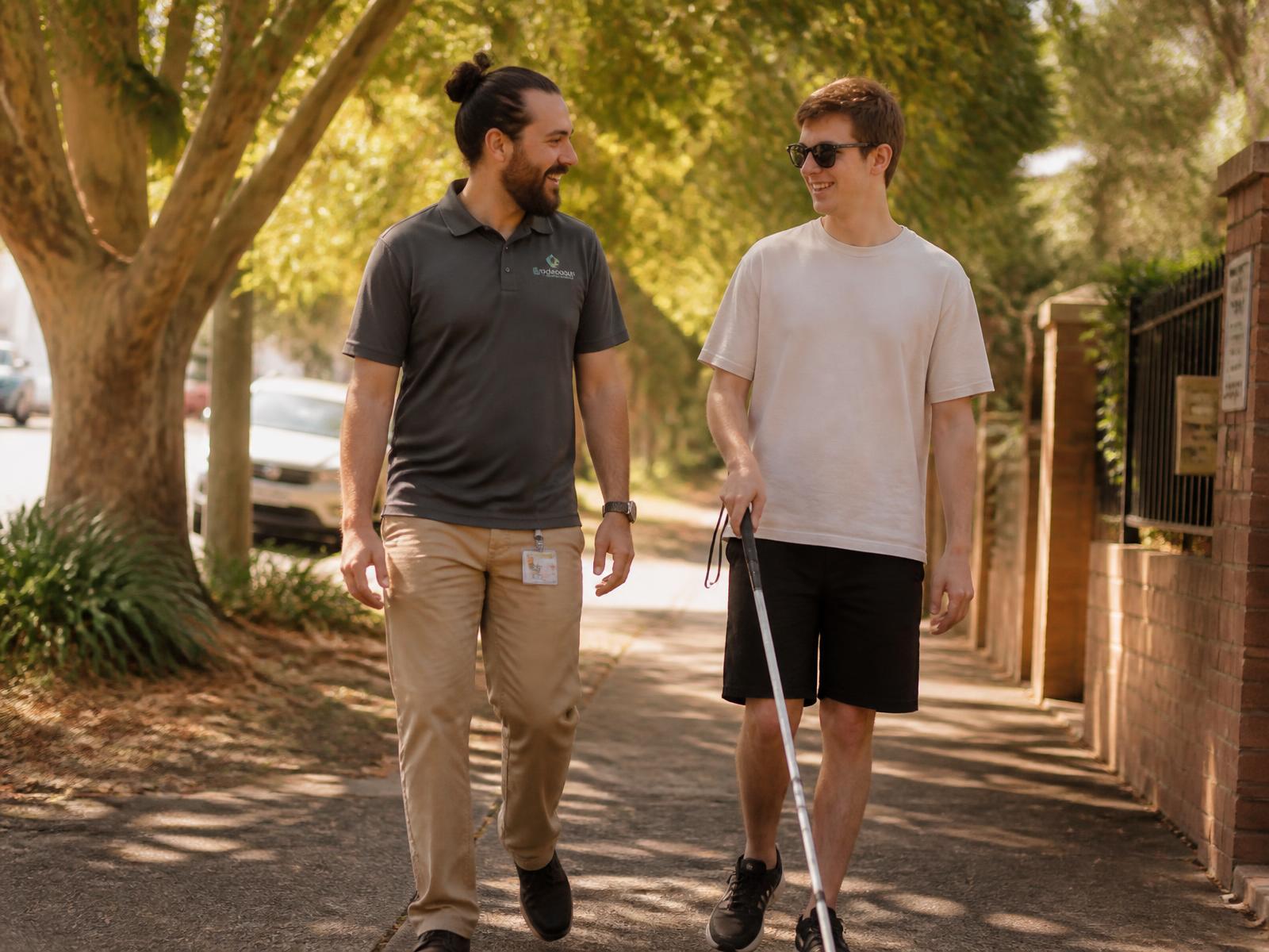 A support worker walking and chatting with a young man who uses a white cane along a tree lined street.